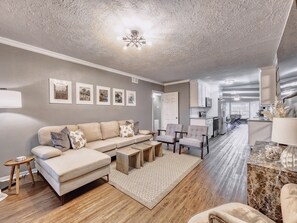 A modern living room with a sectional sofa, a coffee table, and a rug. There is a kitchen area with cabinets and a dining table.