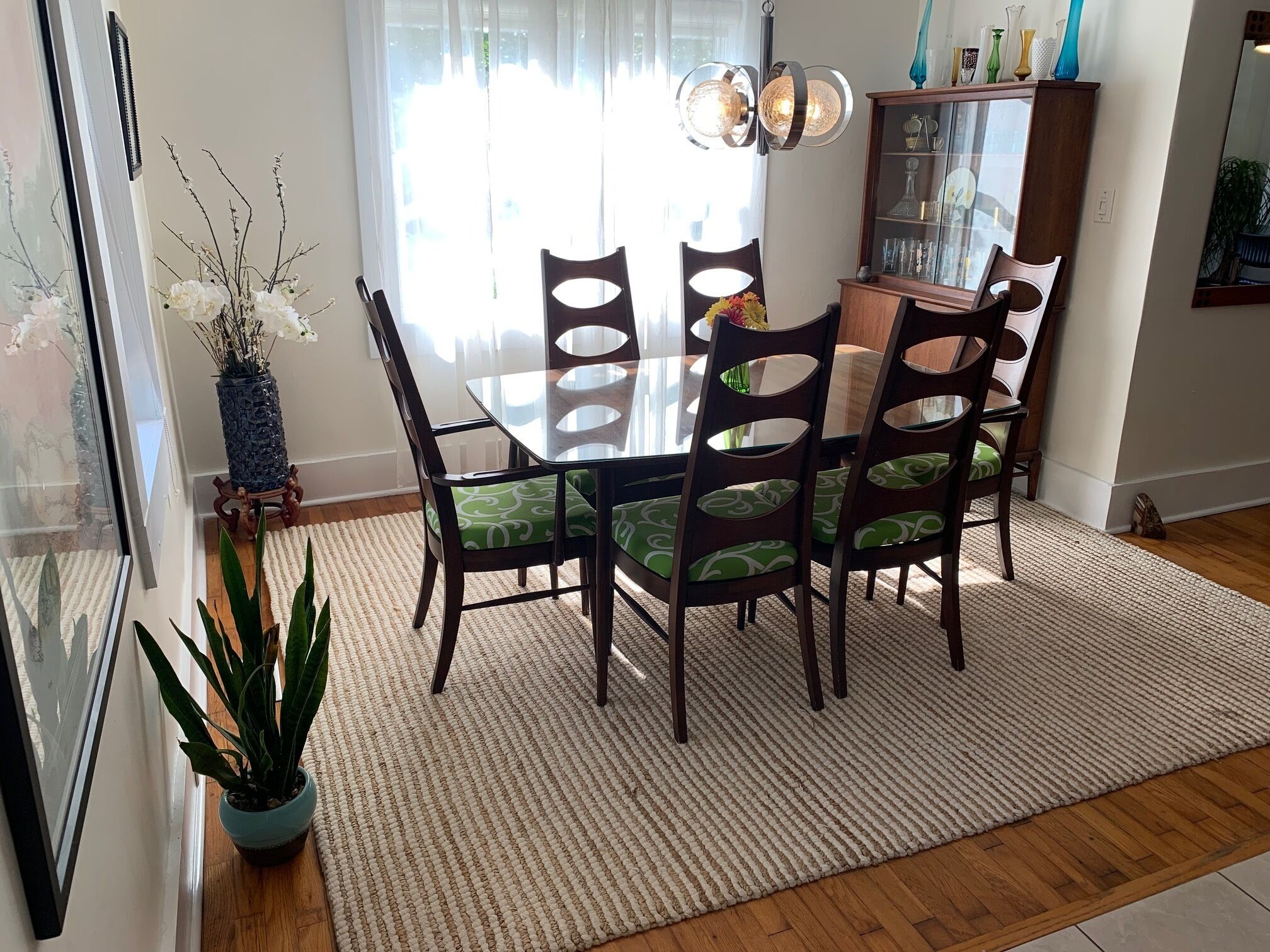 Un coin repas comprenant une table en verre, des chaises en bois, un tapis et un meuble contenant des objets décoratifs.