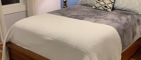 Una cama bien hecha con edredón blanco y manta estampada, una lámpara de noche y una ventana con cortinas.