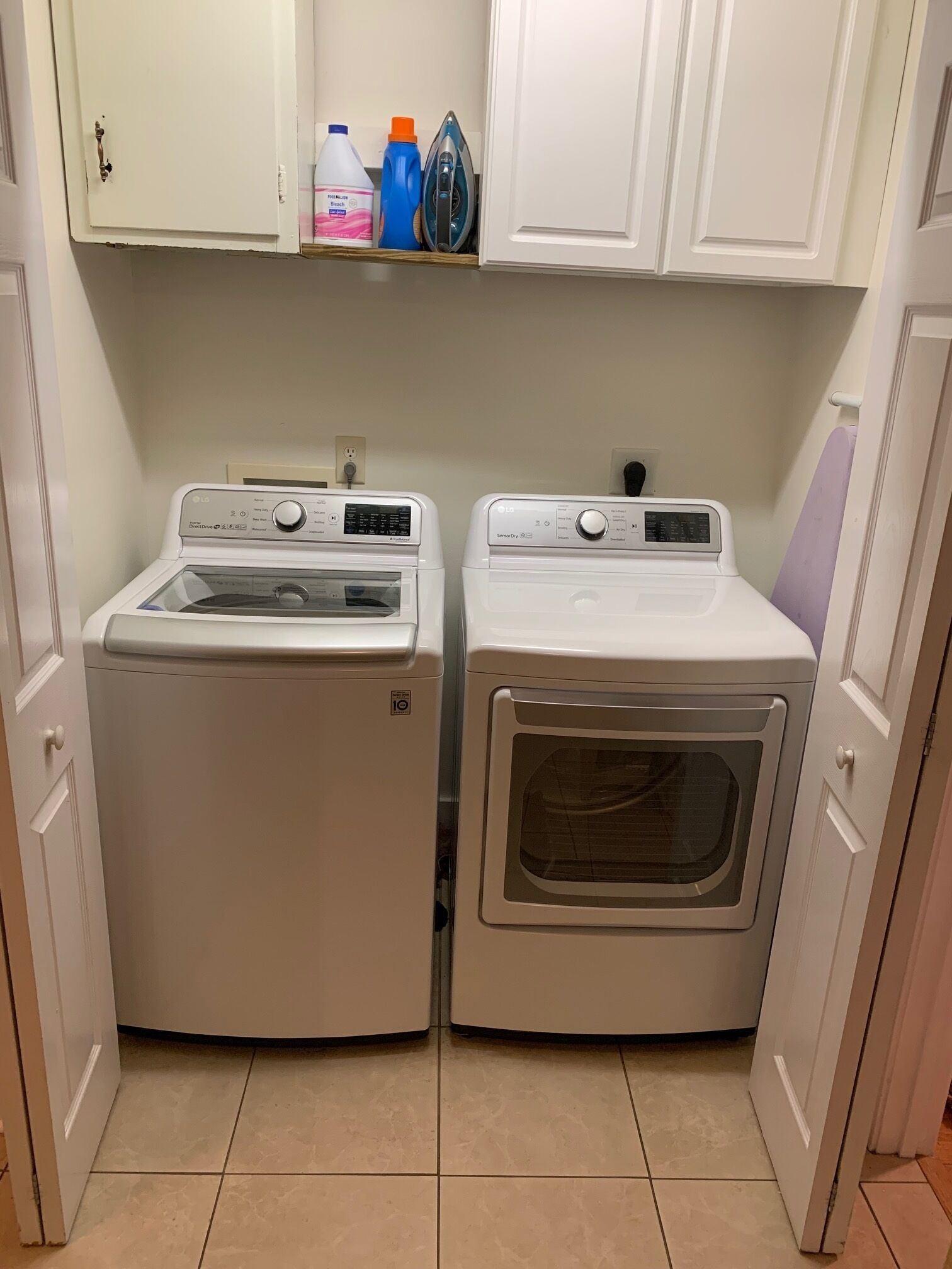 A laundry room with two front-loading washing machines, a shelf with cleaning supplies, and a cabinet above.
