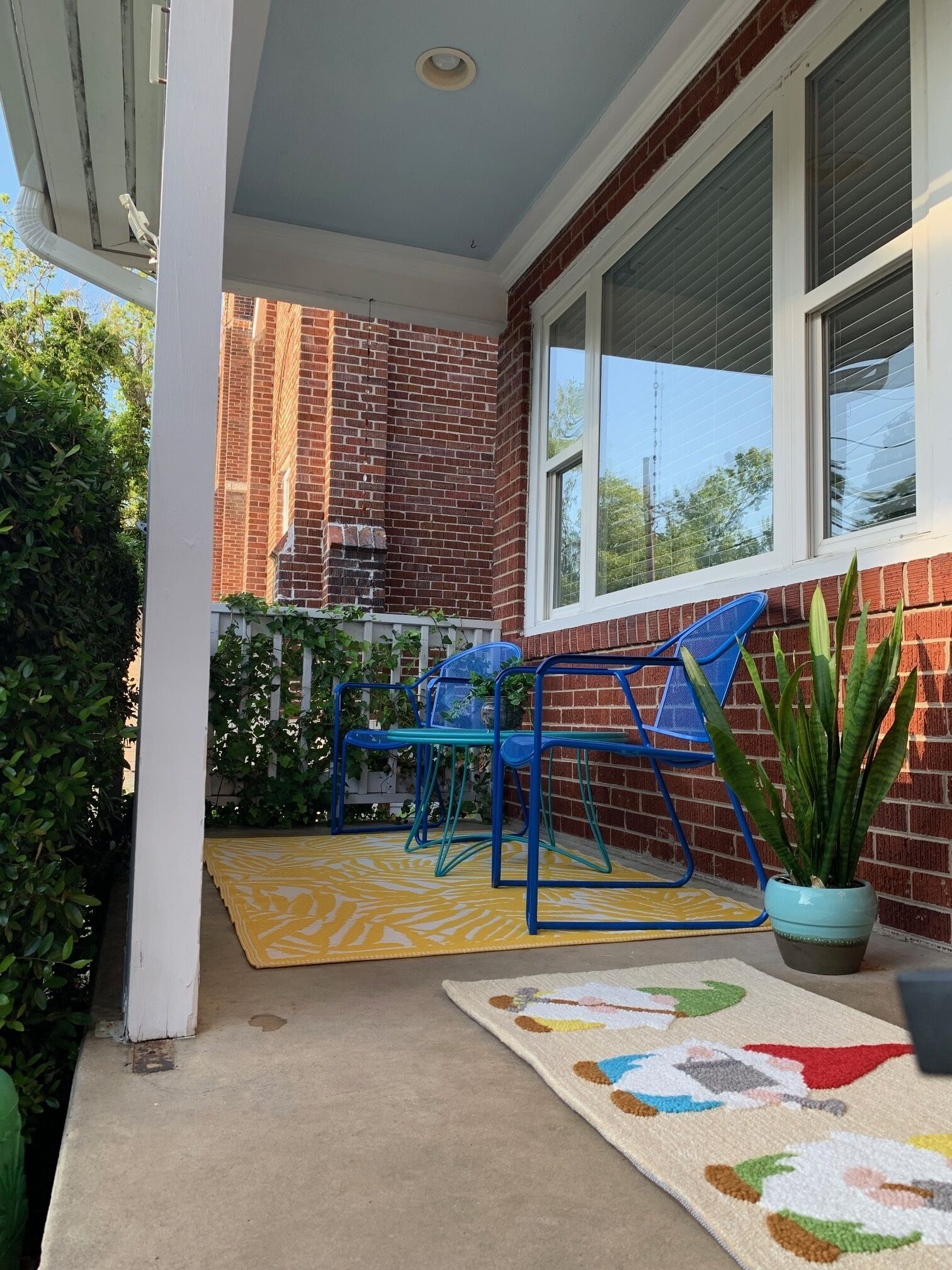 A covered patio with a brick wall, a yellow patterned rug, blue chairs, and a potted plant.