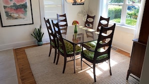 Een eetruimte met een glazen tafel, omringd door zes houten stoelen met groene kussens, een hanglamp en een kunstwerk in een lijst aan de muur.