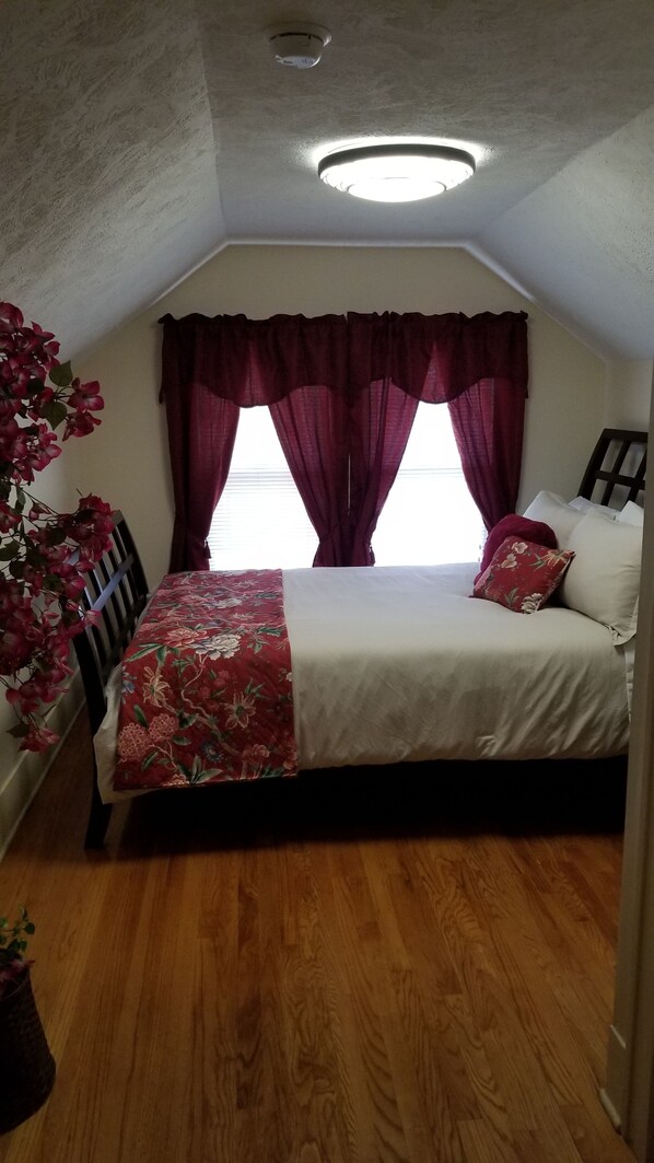 A bedroom with a bed, a window with curtains, a ceiling light, and a wooden floor.