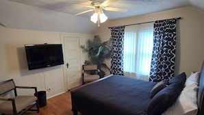 A bedroom with a bed, a chair, a television, a ceiling fan, and a window with patterned curtains.