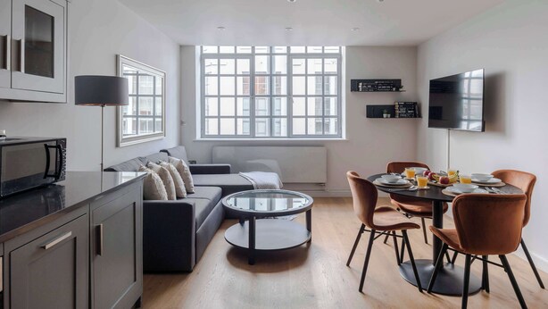 Un espace de vie moderne comprenant un canapé, une table basse ronde, une table à manger avec des chaises, un téléviseur fixé au mur et une fenêtre avec des stores.