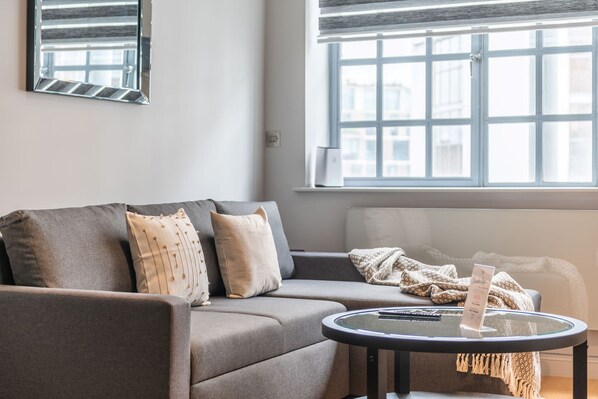 A modern living room with a grey sofa, a glass coffee table, and a window with blinds.
