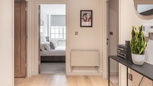 Un couloir menant à une chambre à coucher qui contient un lit, un radiateur, un tableau encadré et une plante en pot.
