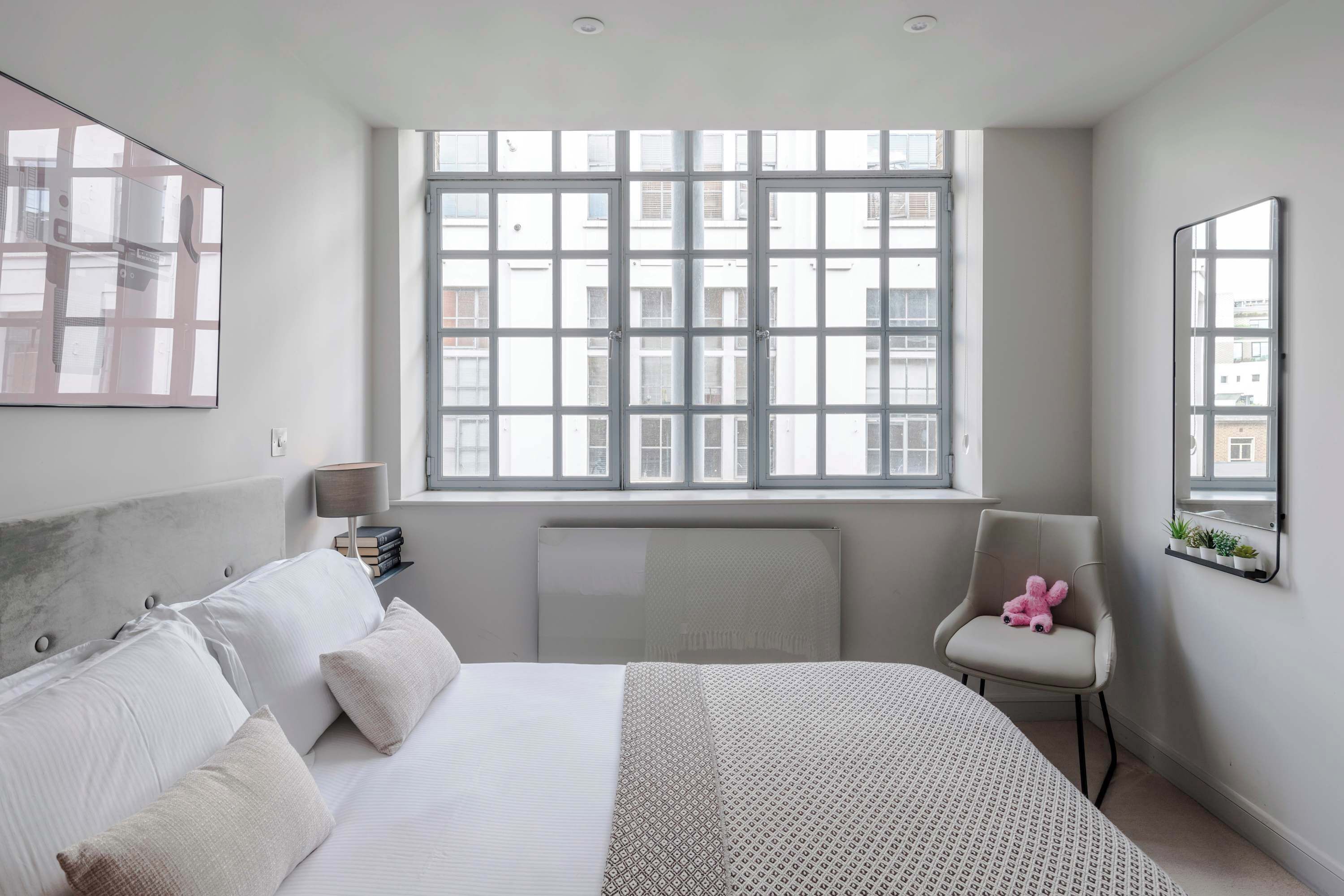 Un dormitorio con un ventanal grande, una cama con ropa de cama blanca, una silla con cojín y una mesita con lámpara.
