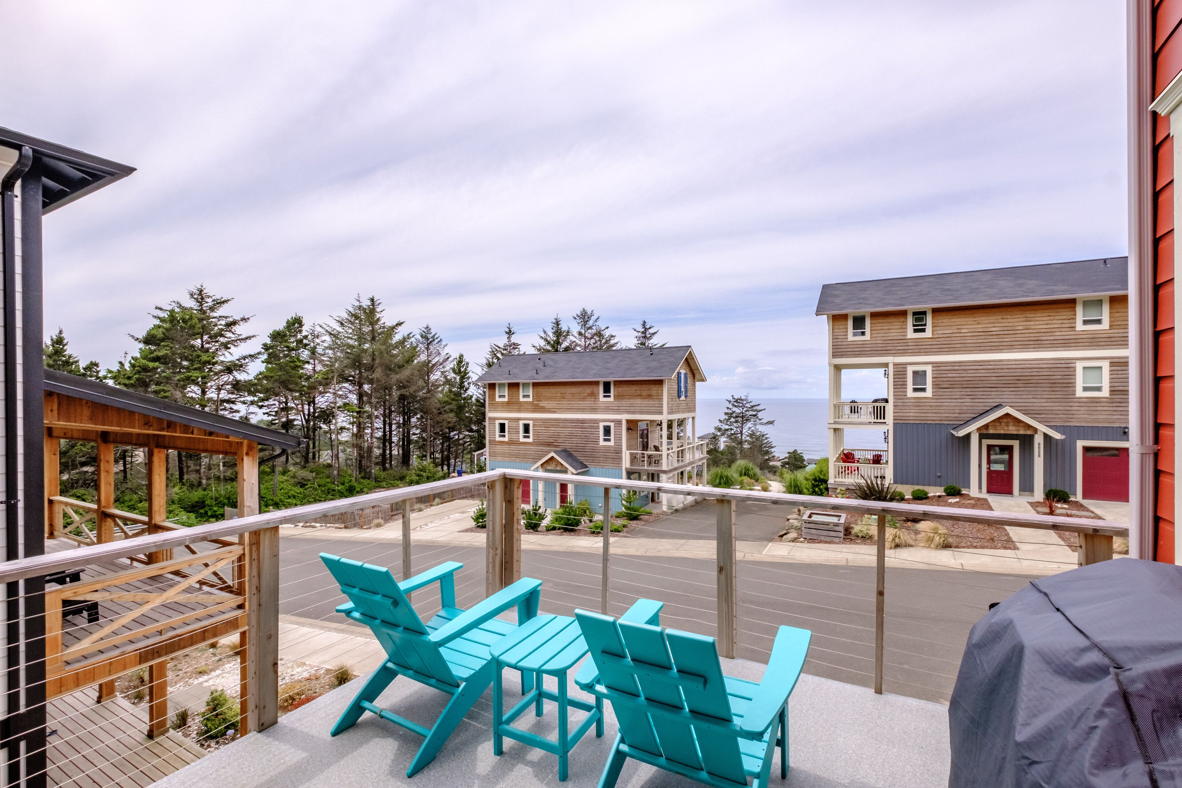 Een balkon met turquoise stoelen en een tafel, met uitzicht op een straat met tweelaags gebouwen en een zeegezicht.