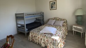 A bedroom with a bunk bed, a single bed with floral bedding, a bedside table with a lamp, and a chair with a cushion. - Maison Traditionnelle en Campagne (Saint-Quentin-de-Caplong)
