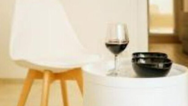 Un intérieur moderne avec une chaise blanche, une table ronde blanche sur laquelle se trouvent un verre à vin et un bol noir, et un tableau accroché au mur.