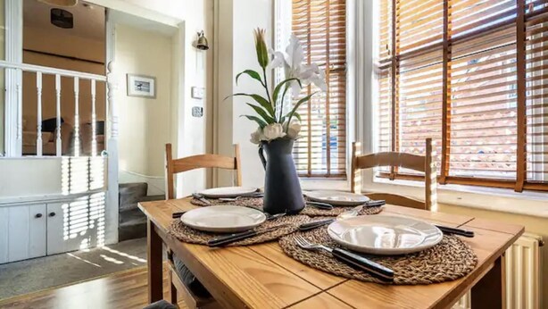 A dining area with a wooden table set for four, a vase with white flowers, and a view through wooden blinds. - The Boat House Sleeps 4 Pet Friendly (Dawlish)