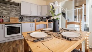 A dining area with a wooden table set for four, surrounded by white cabinets and a kitchen in the background. - The Boat House Sleeps 4 Pet Friendly (Dawlish)