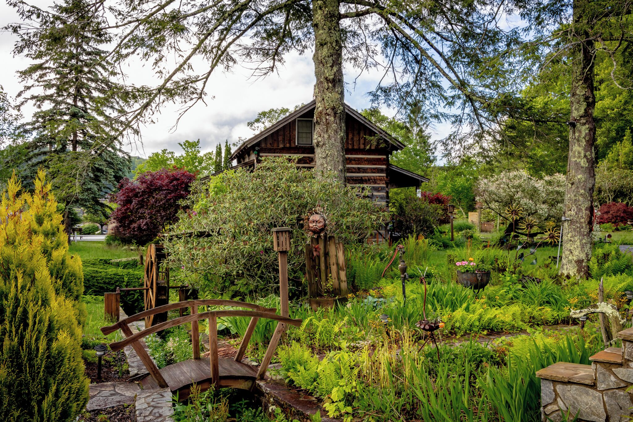 A rustic cabin surrounded by lush greenery, a wooden bridge, and a garden with various plants and a small pond.