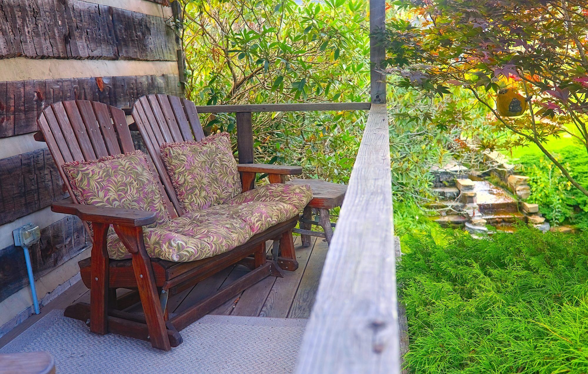 A wooden deck with a bench and a table, surrounded by lush greenery and a small waterfall.