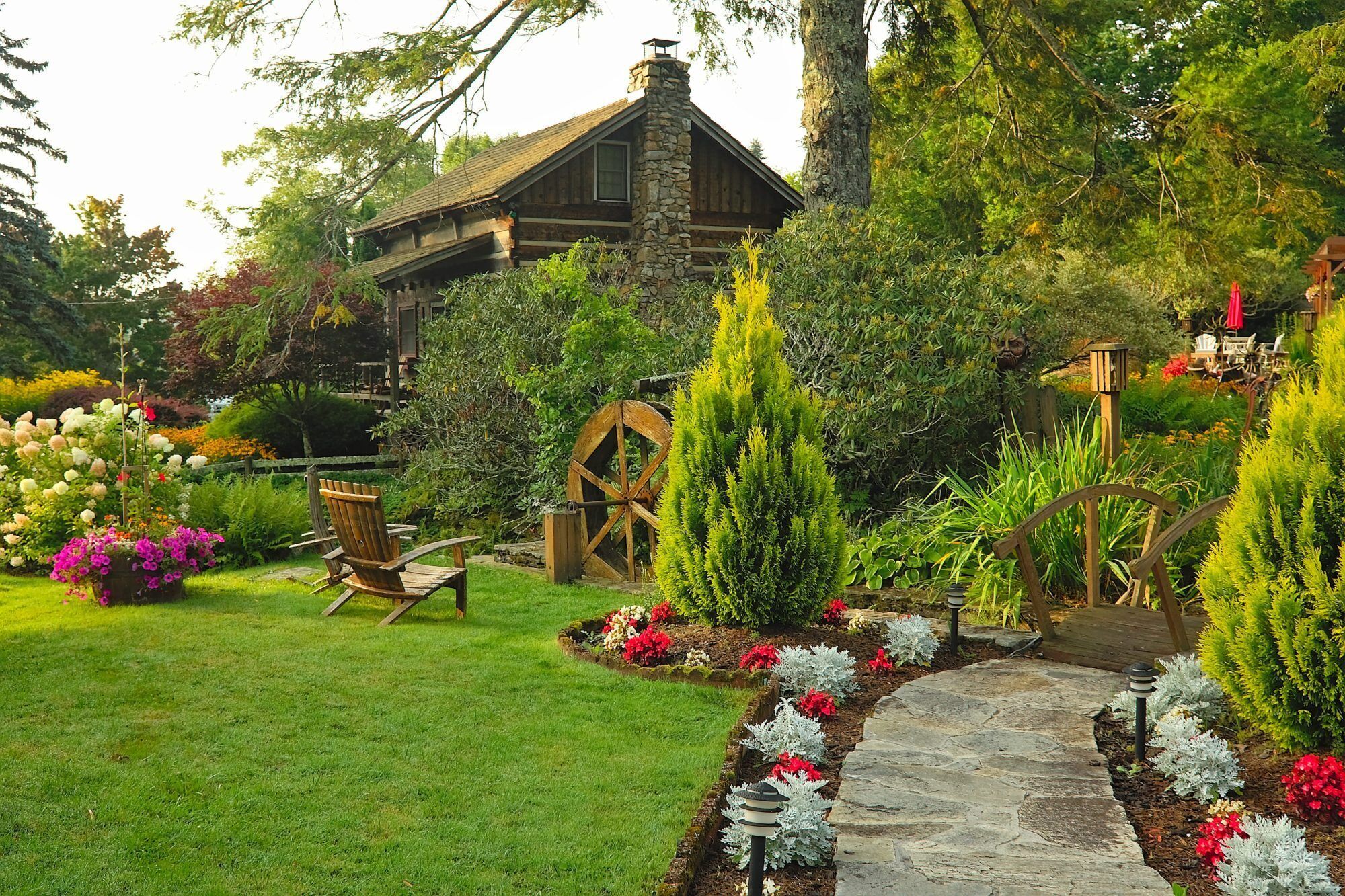 A rustic cabin with a stone chimney, surrounded by a well-maintained garden with a stone pathway, wooden chairs, and a wooden wheelbarrow.