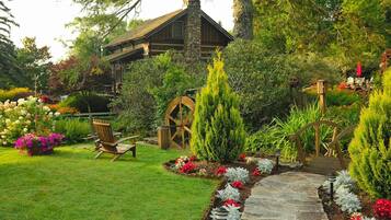 A rustic cabin with a stone chimney, surrounded by a well-maintained garden with a stone pathway, wooden chairs, and a wooden wheelbarrow.