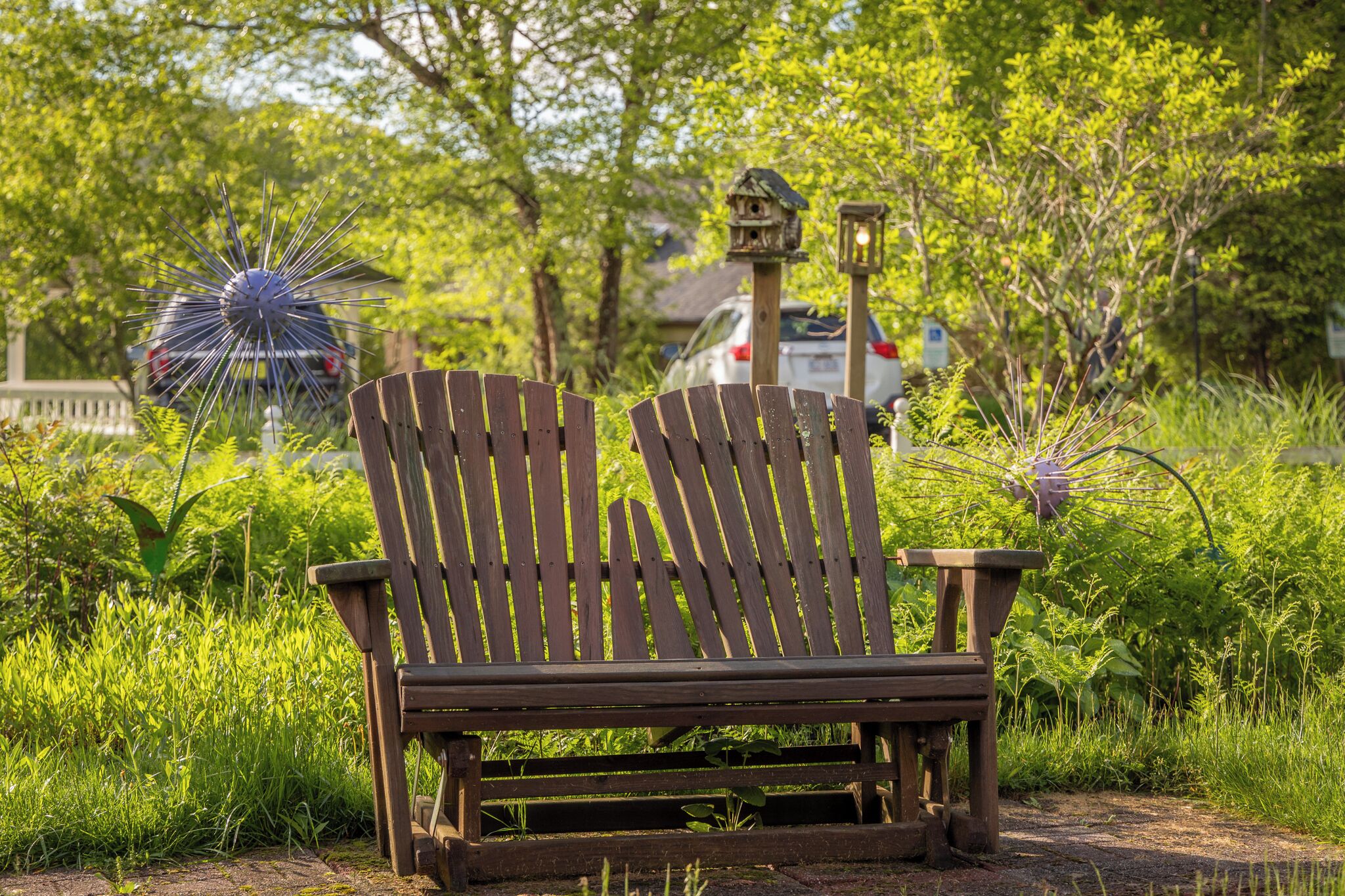Un banc en bois dans un jardin, avec des mangeoires à oiseaux et une voiture en arrière-plan.