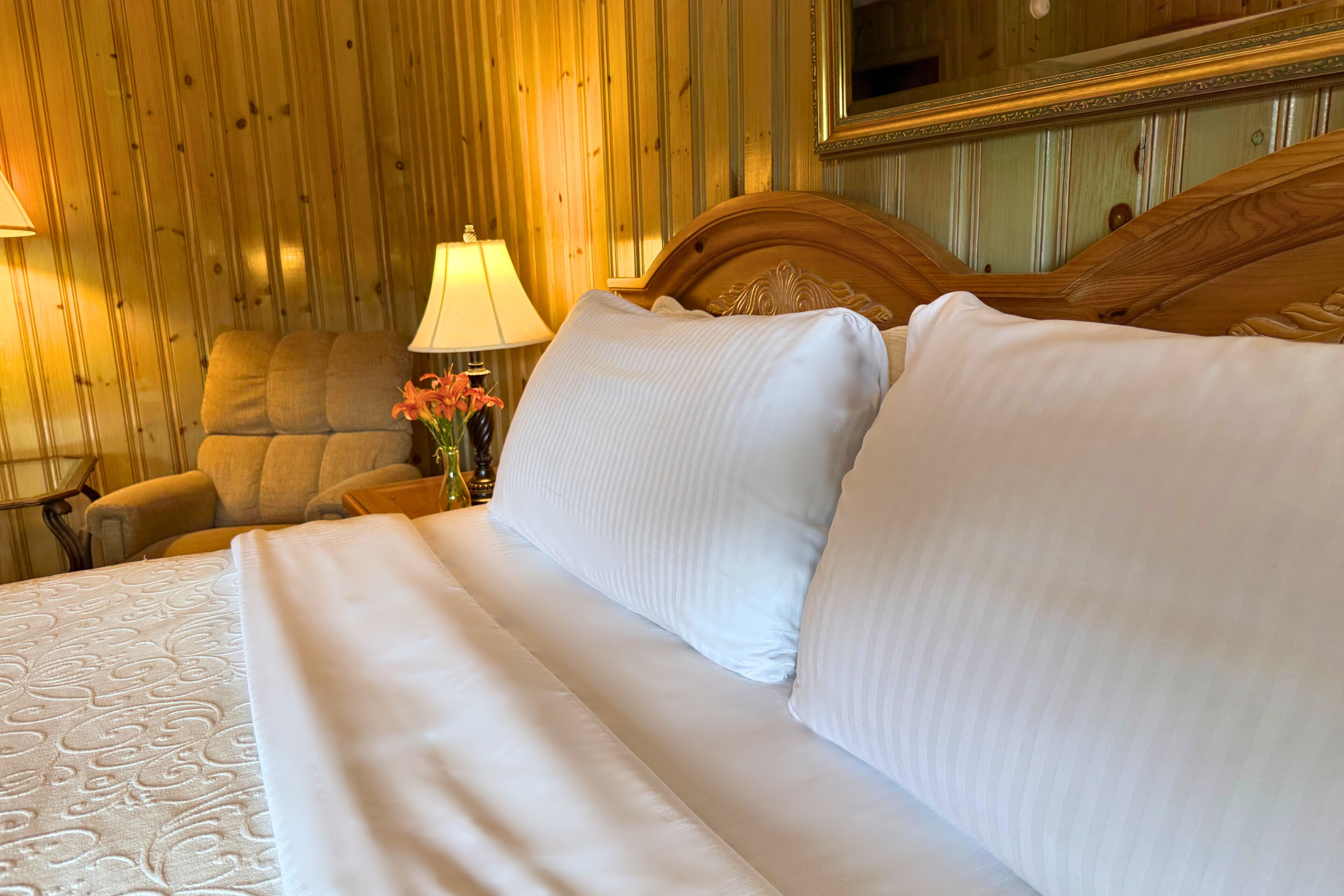A neatly made bed with white linens, a wooden headboard, a lamp, and a chair in a room with wooden paneling.