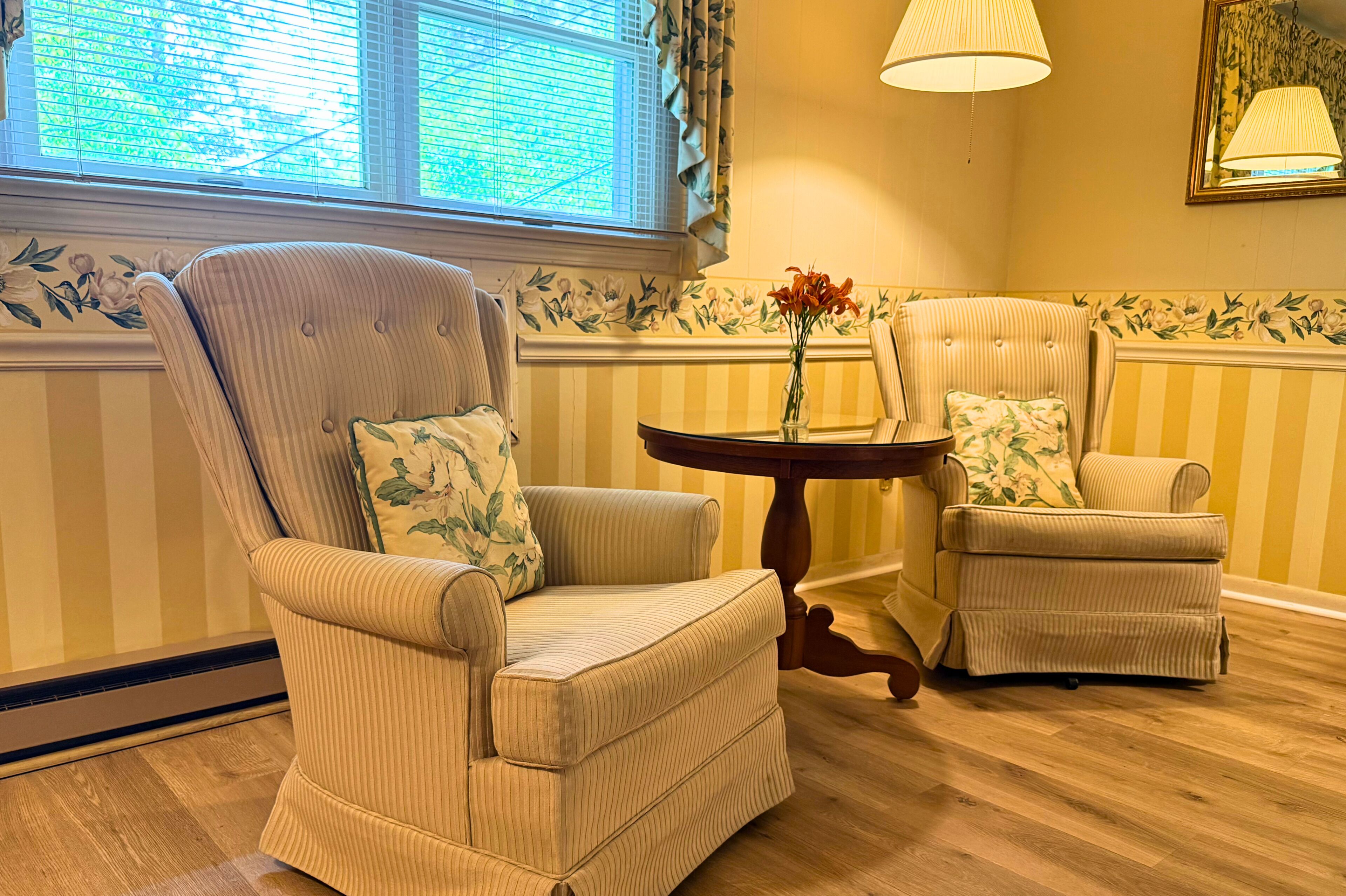 A room with striped wallpaper, two upholstered chairs with floral cushions, a small round table with a vase of flowers, and a window with blinds.