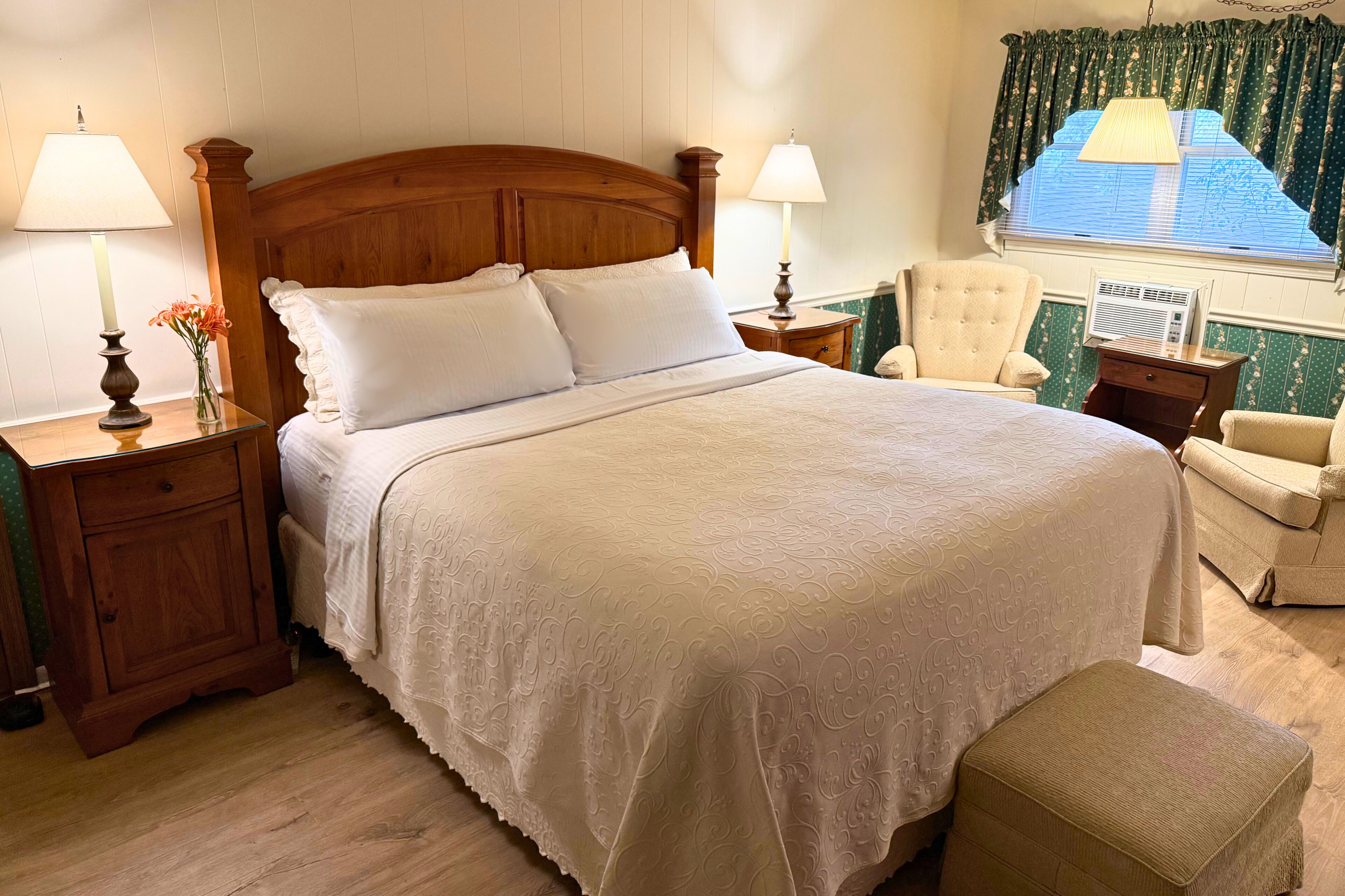 A neatly made bed with a wooden headboard, two bedside tables with lamps, a chair, and a window with curtains.