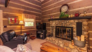 A cozy log cabin interior with a stone fireplace, leather sofa, and a television. - Azalea Garden Inn (Blowing Rock)