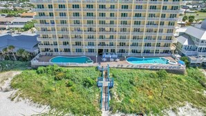 A multi-story hotel with a pool area, a staircase leading to the pool, and a beach in the foreground.