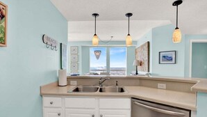 A kitchen with a double sink, white cabinets, a dishwasher, pendant lights, and a view of the city through a window.