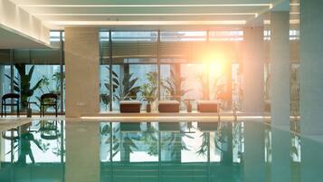 A modern indoor pool area with glass walls, lounge chairs, and potted plants.
