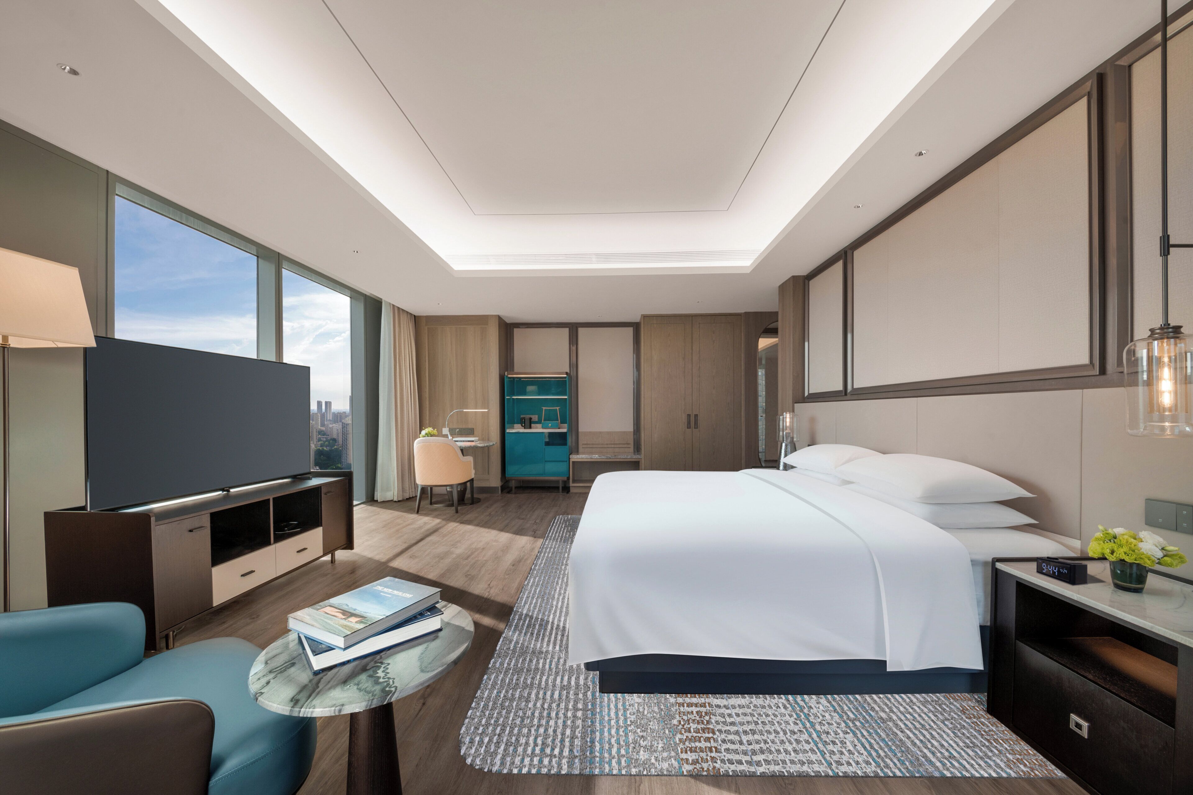 A modern hotel room with a large bed, a TV, a desk, and a view of the city.
