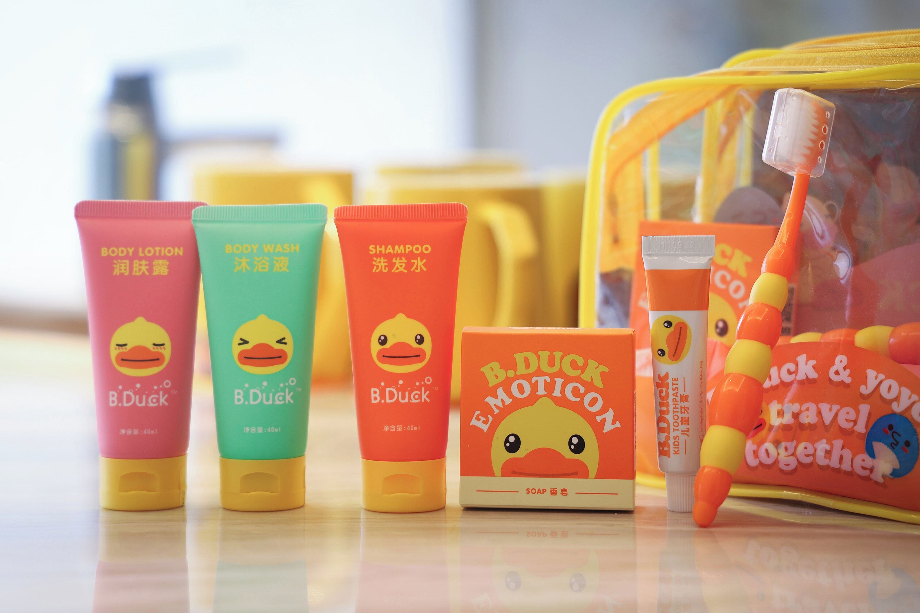 Una colección de productos de tocador de la marca B. Duck, incluyendo loción corporal, jabón de ducha, champú y pasta de dientes, sobre una superficie reflectante.