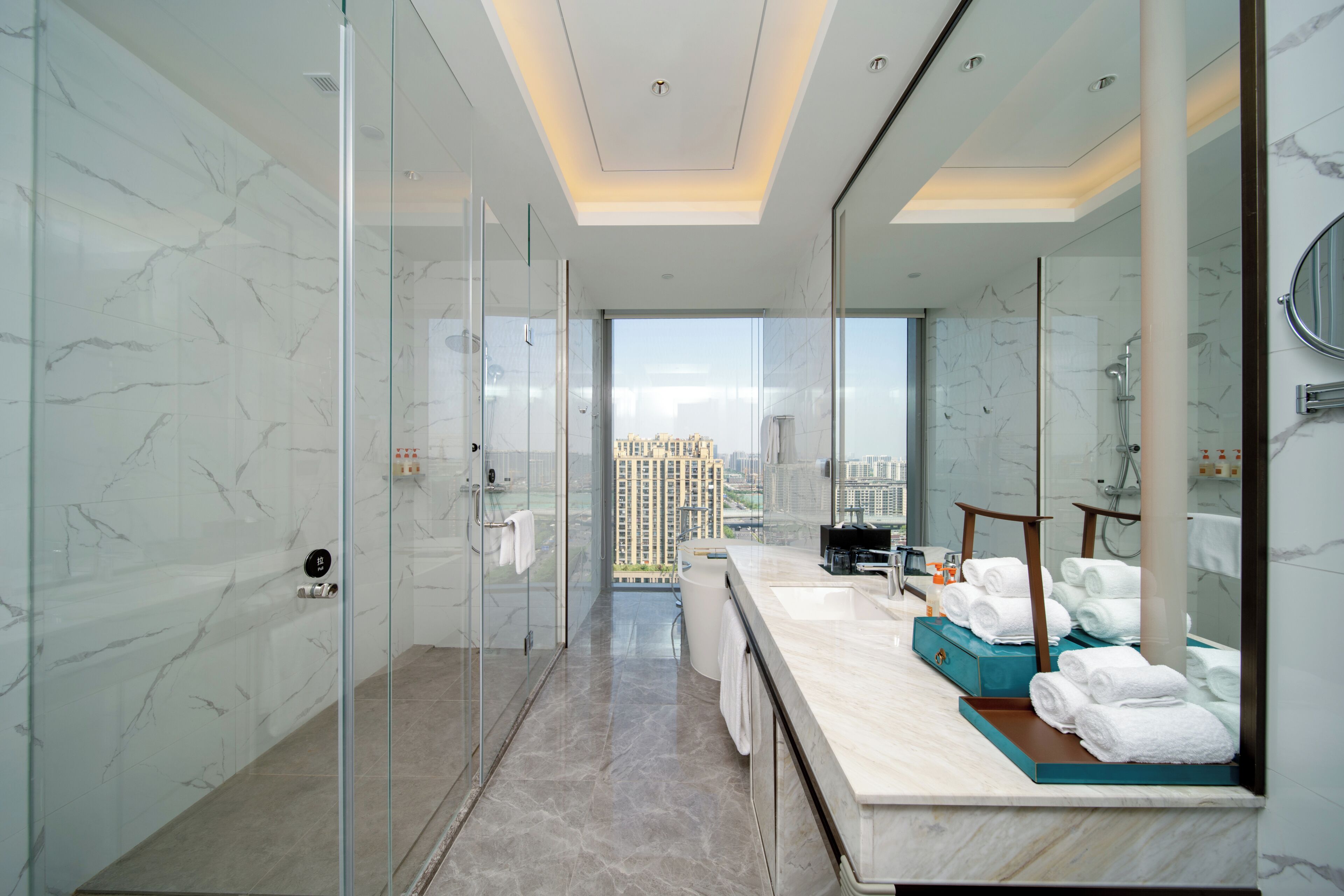 Un baño moderno con cabina de ducha de vidrio, un espejo grande y vistas a un paisaje urbano.