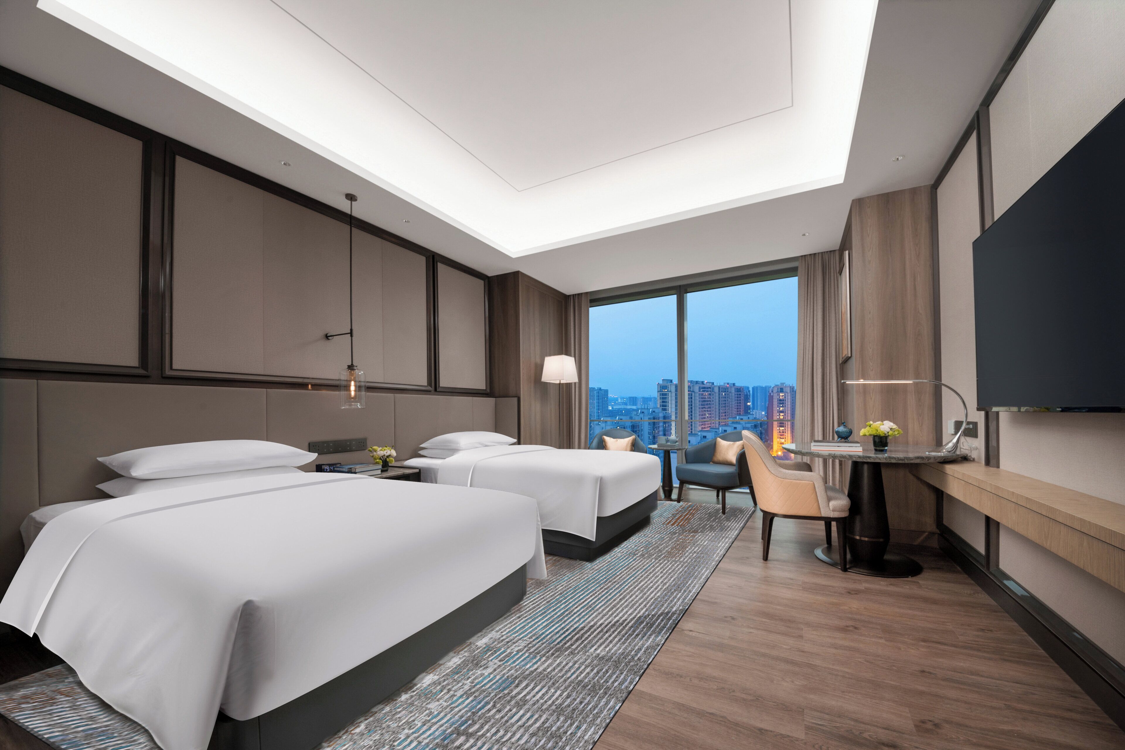 A modern hotel room with two beds, a desk, a chair, and a large TV.