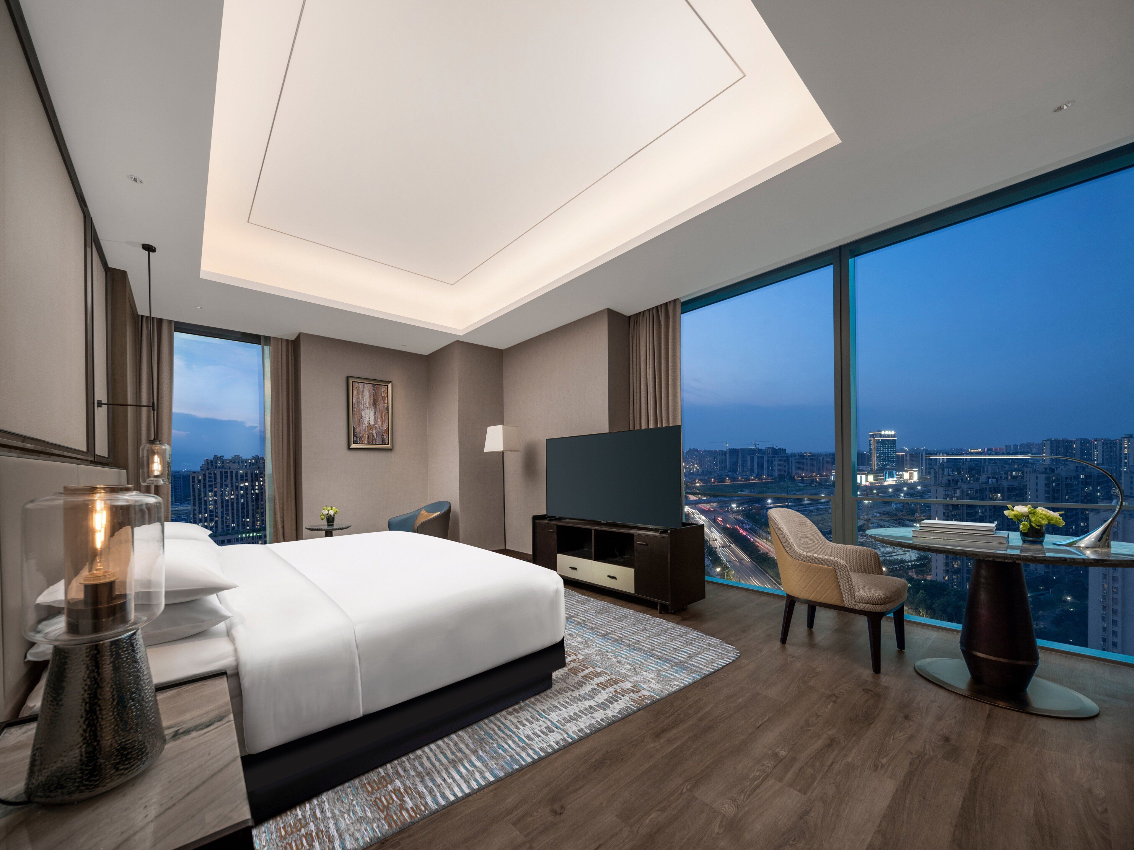 Habitación de hotel moderna con una cama grande, televisión, escritorio y balcón con vistas a la ciudad.