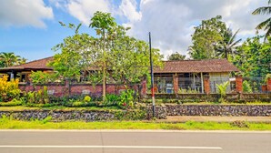 A two-story building with a tiled roof, surrounded by lush greenery and a stone wall. - Hotel O Sekar Jagat Near Pantai Baluk Rening (Negara)