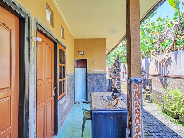 A narrow corridor with a wooden door, a small room with a toilet, and a view of a garden with banana plants. - Hotel O Sekar Jagat Near Pantai Baluk Rening (Negara)