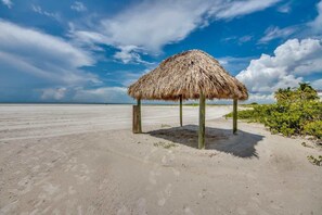 A beach with a thatched-roof structure, sandy beach, and clear sky. - Pelican Watch #105 Beautiful Beachfront Condo (Fort Myers Beach)