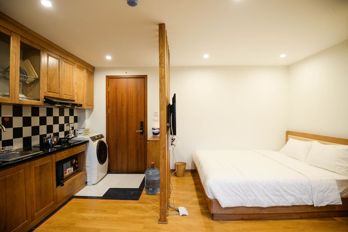 A compact hotel room with a wooden bed, a washing machine, a kitchenette, and a wooden wardrobe.