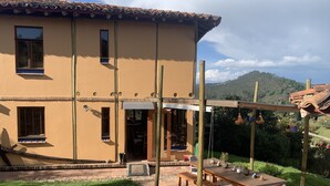 A two-story house with a patio area featuring a wooden table and chairs, a small garden, and a view of the surrounding hills. - El Respiro de La Colina (Tuta)