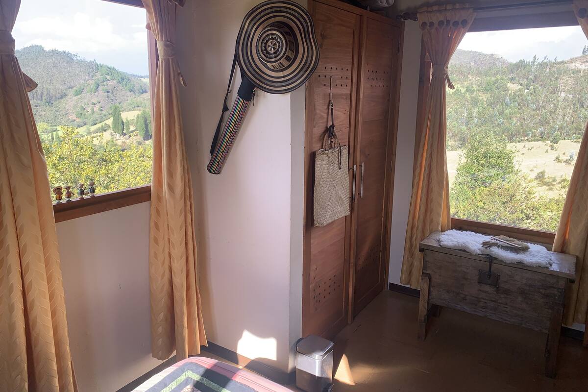 A room with a bed, a wardrobe, a window with curtains, and a view of a hilly landscape.