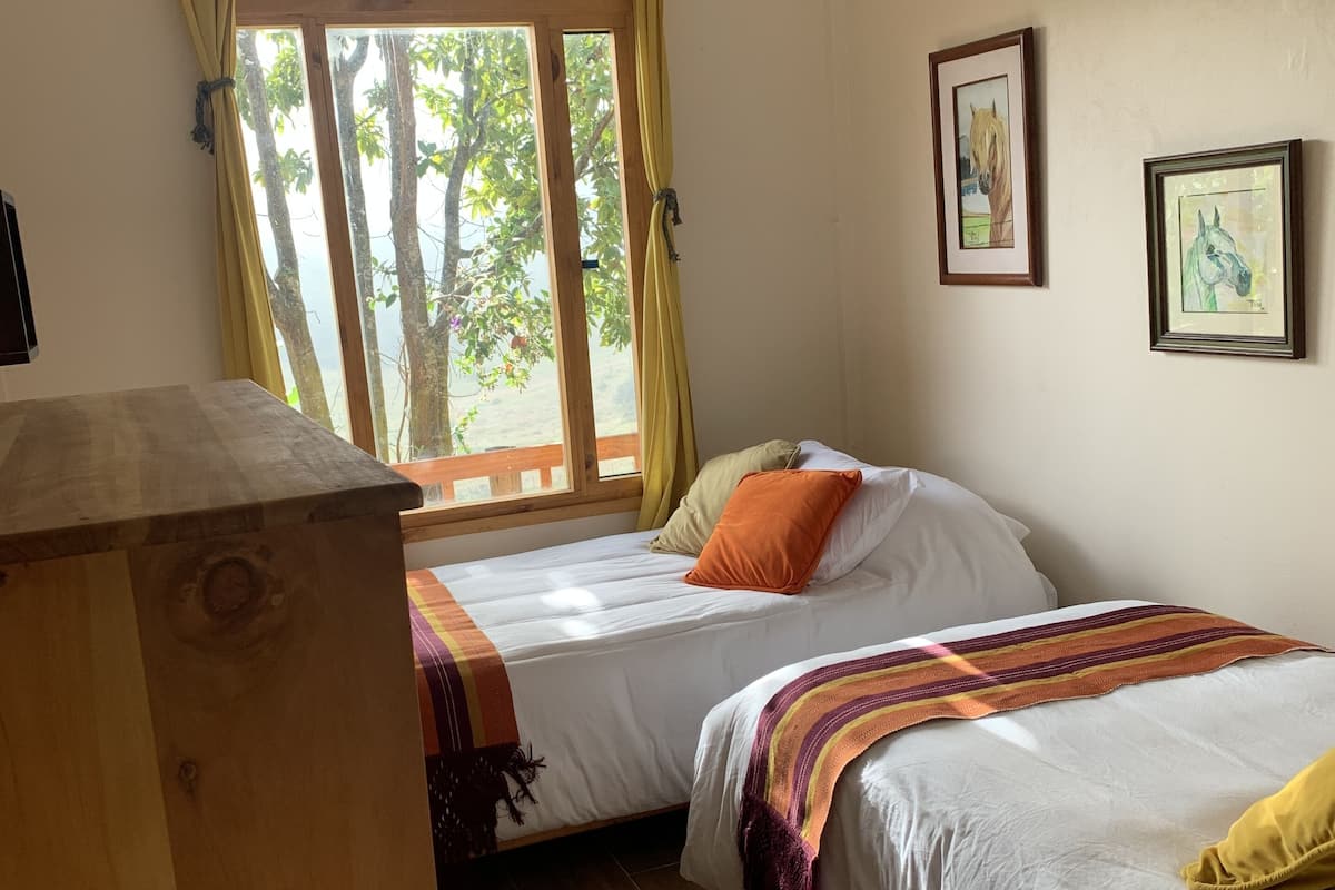 A bedroom with two beds, wooden flooring, a window with curtains, and framed artwork on the wall.