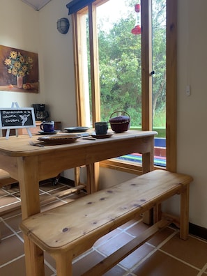 Una mesa de madera con un letrero de pizarra, una taza azul, una cesta y un banco de madera en una habitación con ventanas amplias y vistas a zonas verdes.