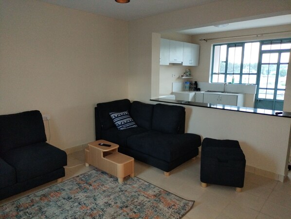 Ein Wohnzimmer mit einer Couch, Hocker und einem kleinen Holzschemel. Es gibt eine Küchenzeile mit Schränken und einem Fenster.