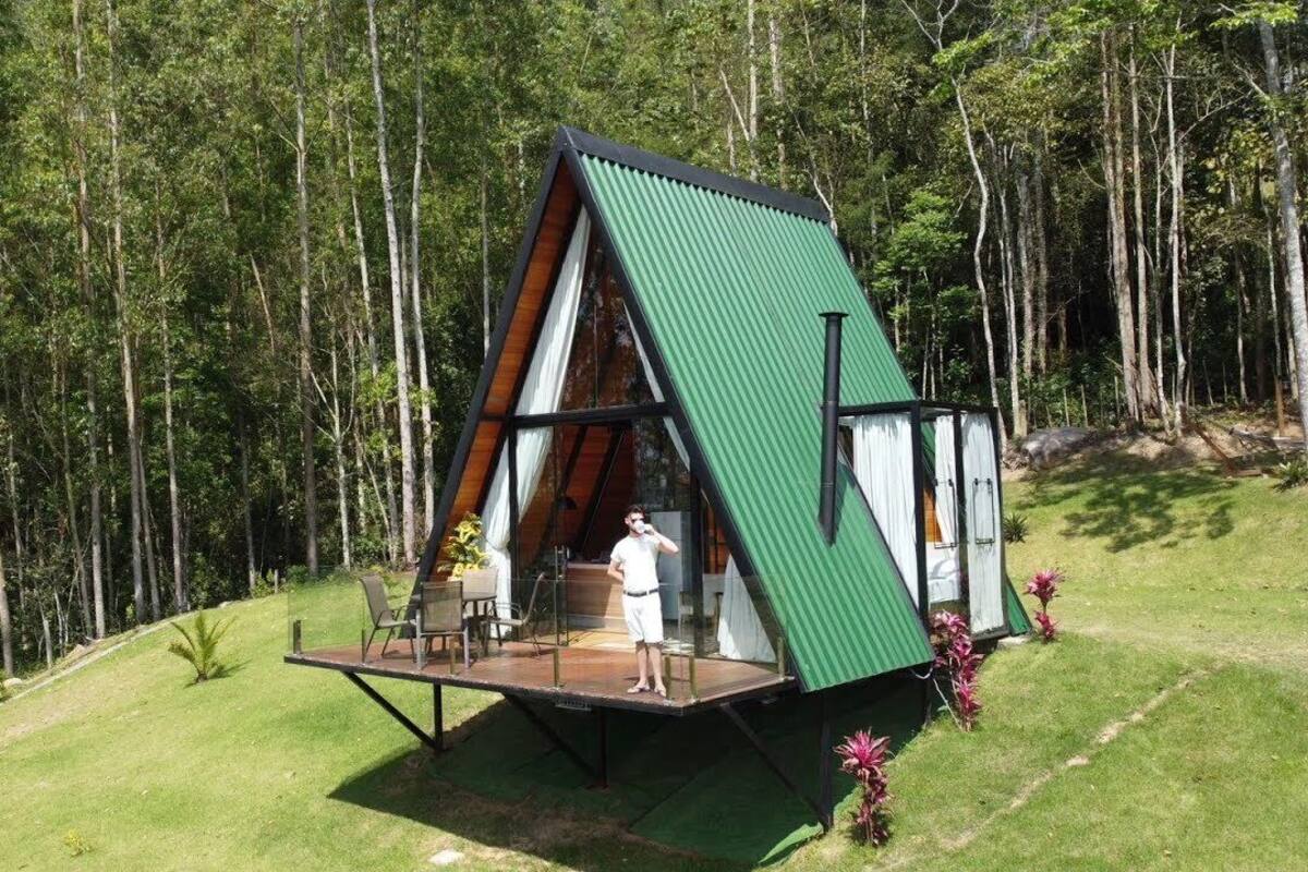 Eine moderne, dreieckige Holzhütte mit grüner Dachbegrünung, gelegen auf einem grasbewachsen Hügel, umgeben von Bäumen.