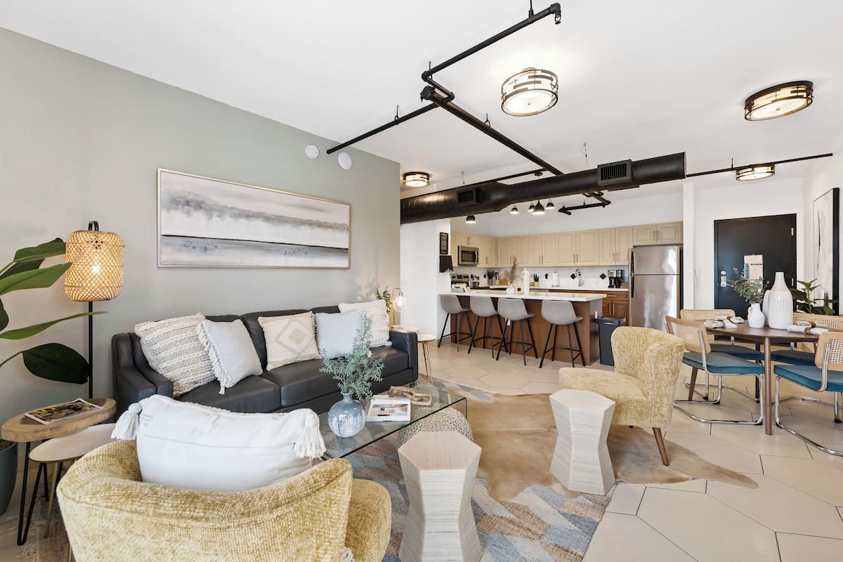 A modern living room with a sofa, coffee table, and chairs, a dining area with a table and chairs, and a kitchen in the background.
