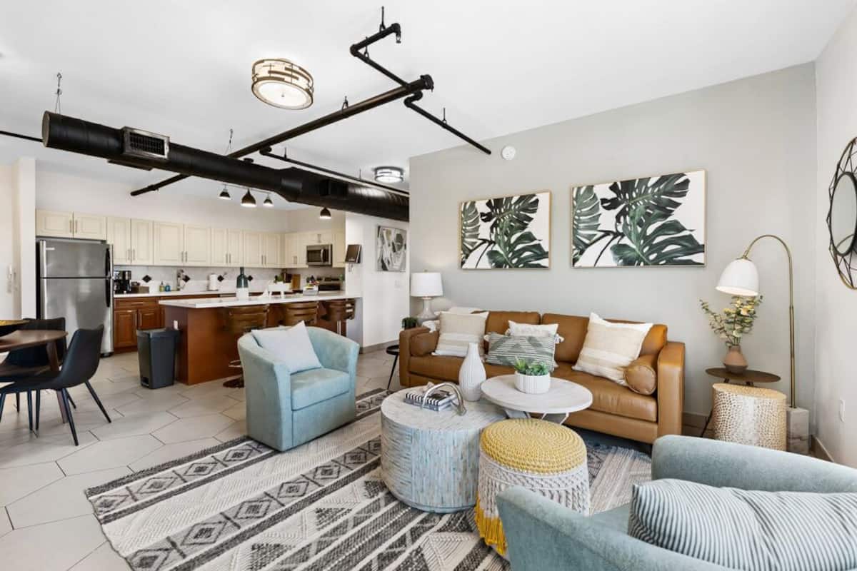 A modern living room with a sofa, coffee table, and ottoman, a kitchen in the background, and artwork on the walls.