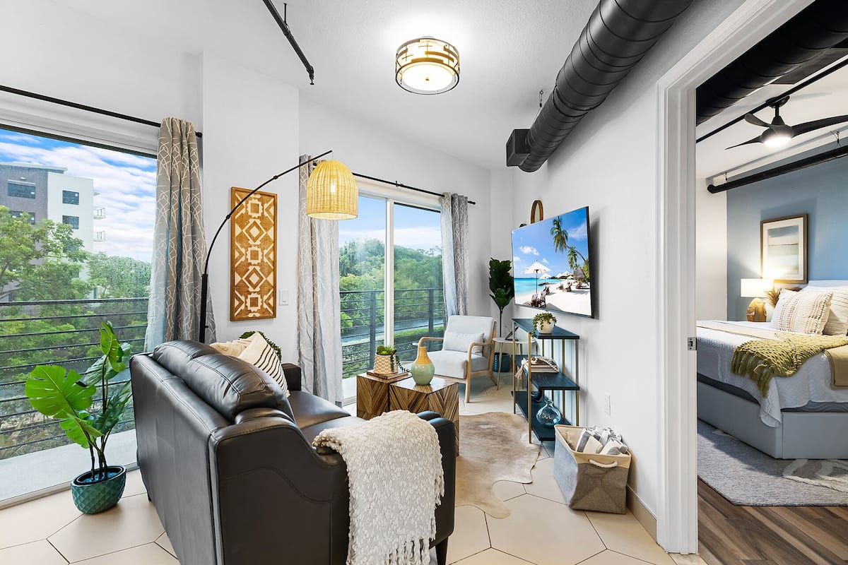 A modern living room with a leather sofa, a dining area with a table and chairs, a large TV, and a balcony with a view of buildings and trees.