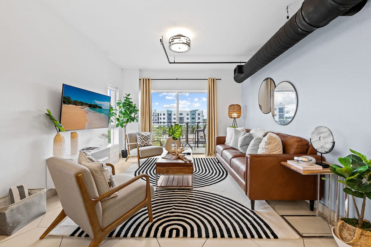 A modern living room with a sofa, armchair, coffee table, and a large black and white striped rug. There is a TV mounted on the wall, a plant, and a window with a view of buildings.