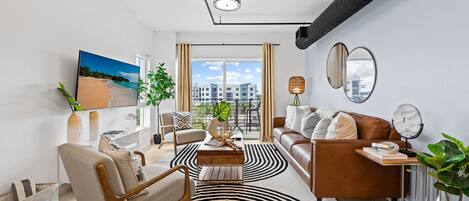 Ett modernt vardagsrum med en soffa, fåtölj, soffbord och en stor svartvit randig matta. På väggen sitter en tv, och det finns även en växt och ett fönster med utsikt över byggnader.