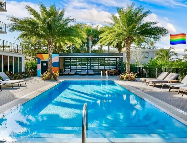 Piscina con sillones de descanso, palmeras y un edificio moderno con paneles de colores.